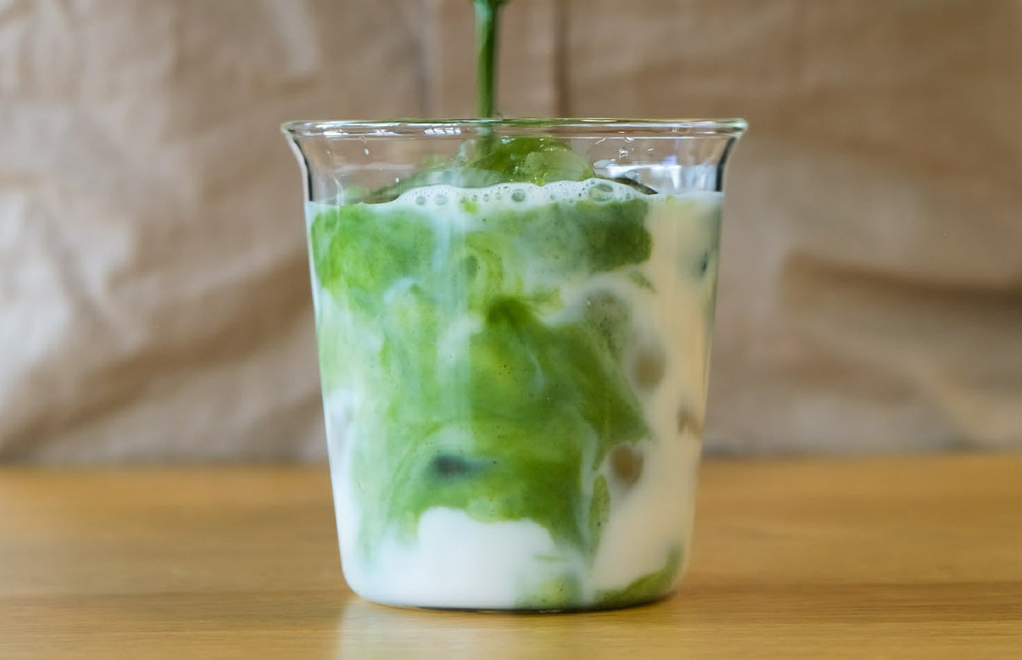 Green matcha liquid being poured into a glass with white foam on a wooden surface